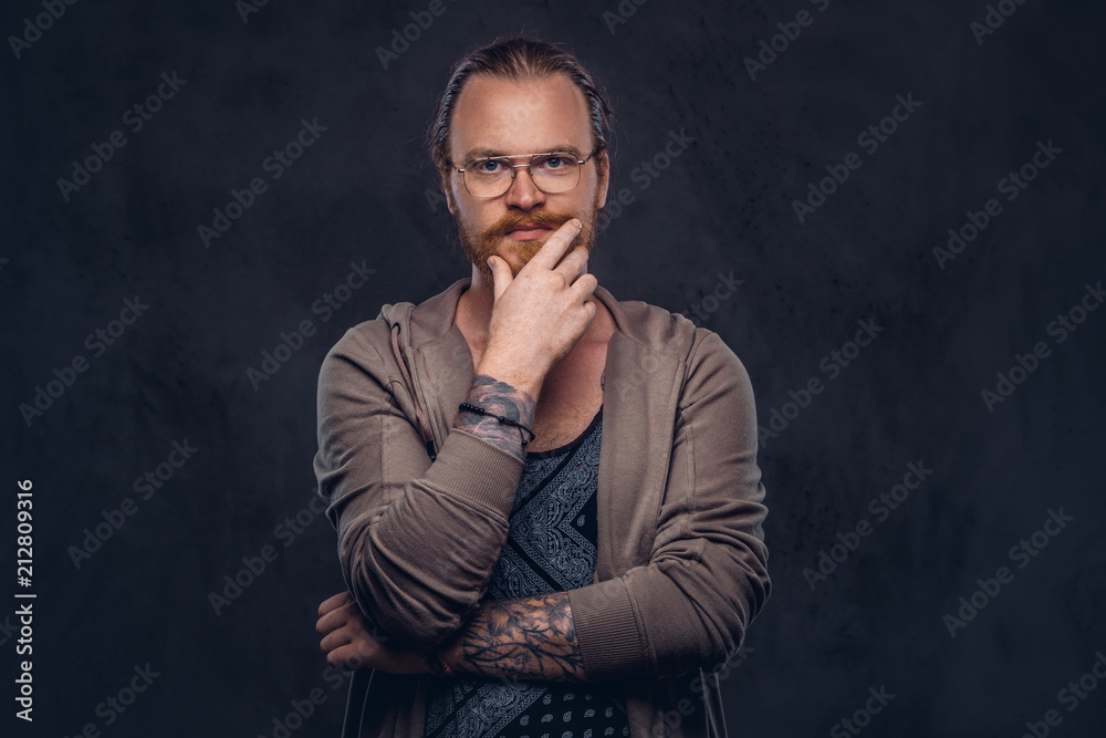 Smart pensive redhead hipster with full beard and glasses dressed in casual clothes, poses with hand on chin in a studio. Isolated on a dark background. Smart pensive redhead hipster with full beard and glasses dressed in casual clothes, poses with hand on chin in a studio. Isolated on a dark background.