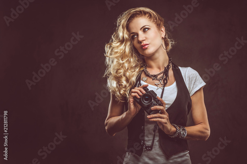 Sensual curly blonde girl photographer dressed in a white t-shirt and waistcoat wears a lot of accessories and wristwatch, posing with a camera at a studio. Isolated on a dark textured background.