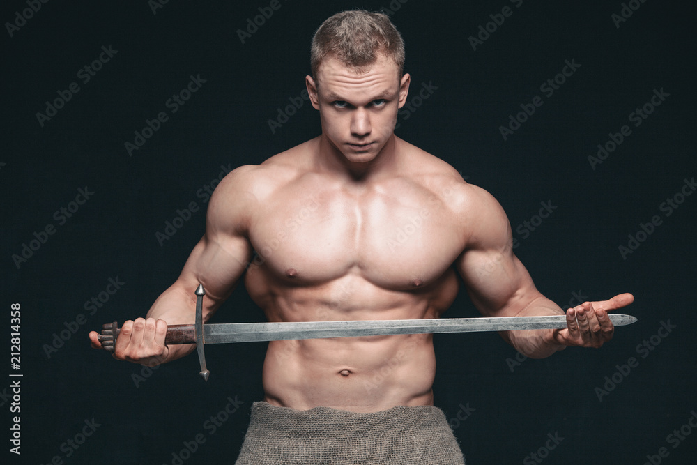 Bodybuilder man posing with a sword isolated on black background. Serious shirtless man demonstrating his mascular body Bodybuilder man posing with a sword isolated on black background. Serious shirtless man demonstrating his mascular body
