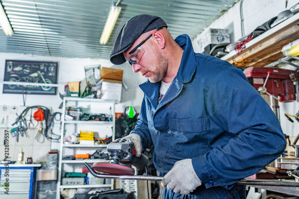 A young man welder in a blue T-shirt, goggles and construction gloves processes metal an angle grinder in the garage, in the background a lot of tools