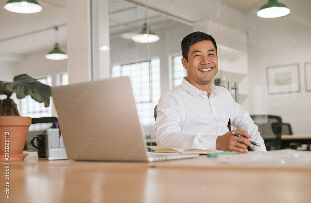 Smiling young Asian businessman working alone at his office desk