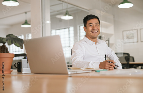 Smiling young Asian businessman working alone at his office desk