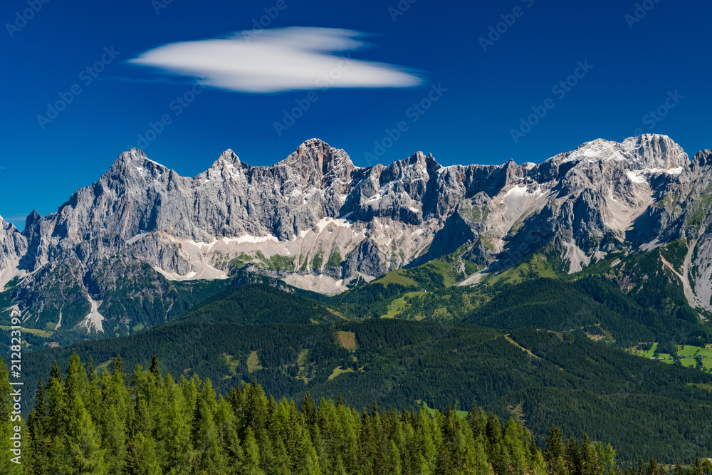 Hohe Dachstein mountain range in Austria with green trees in the foreground and a white cloud on blue sky