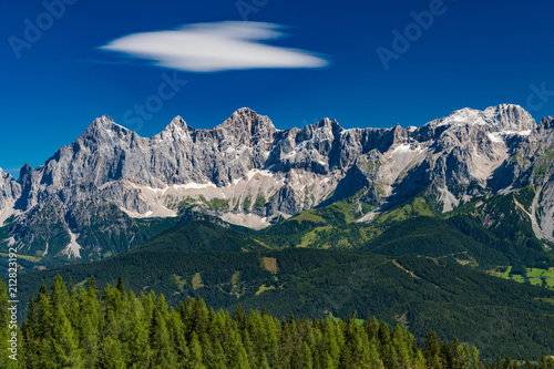 Hohe Dachstein mountain range in Austria with green trees in the foreground and a white cloud on blue sky