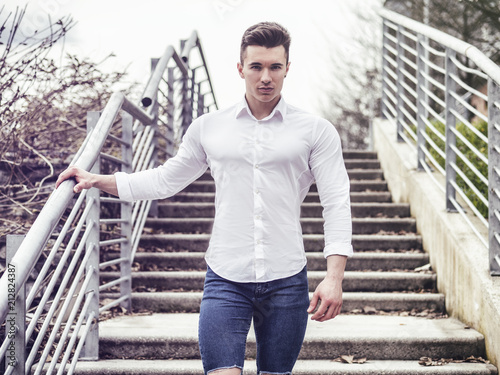 One handsome young man in urban setting in European city, standing, wearing white shirte and jeans