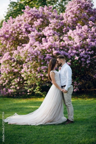 Romantic tender wedding couple of groom and bride hugging near the big purple flower bush in the garden.