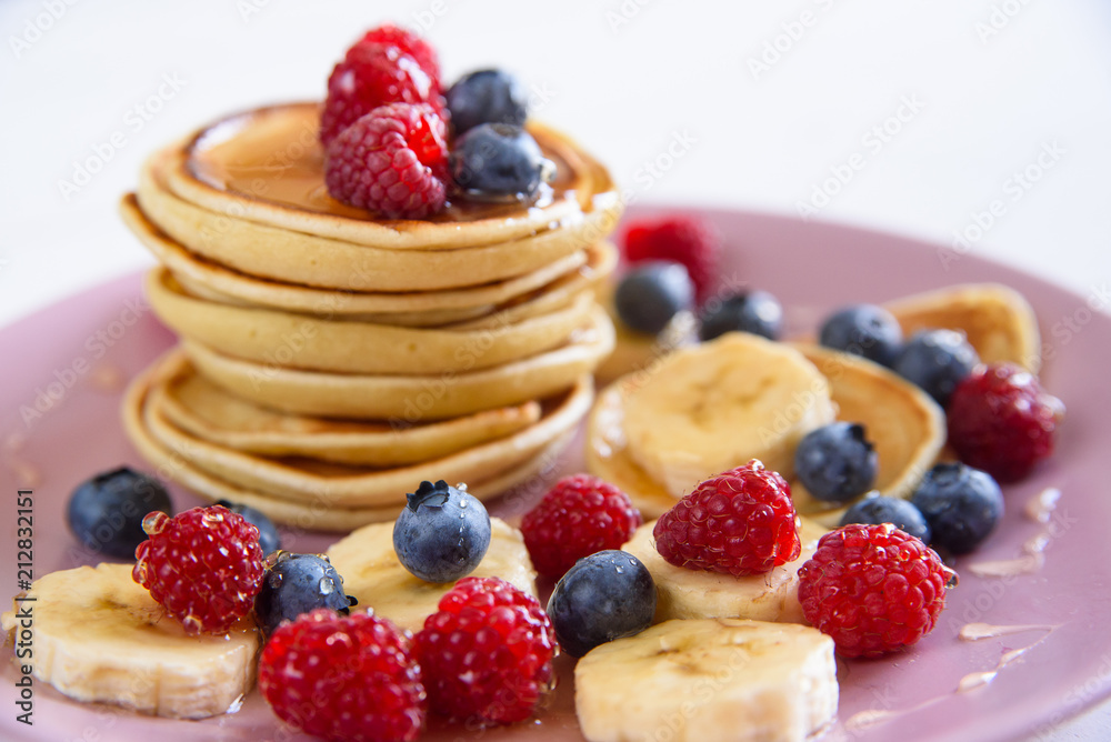 Delicious homemade pancakes with fresh berries in a violet plate on a white background. A tasty and healthy breakfast of pancakes with raspberries, blueberries and honey. Delicious homemade pancakes with fresh berries in a violet plate on a white background. A tasty and healthy breakfast of pancakes with raspberries, blueberries and honey.
