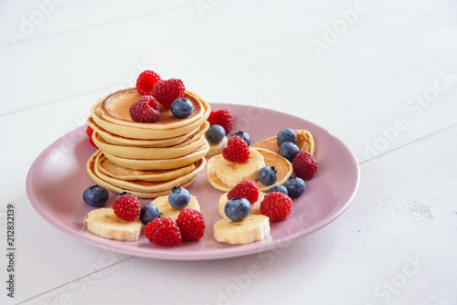 Delicious homemade pancakes with fresh berries in a violet plate on a white background. A tasty and healthy breakfast of pancakes with raspberries, blueberries and bananas.