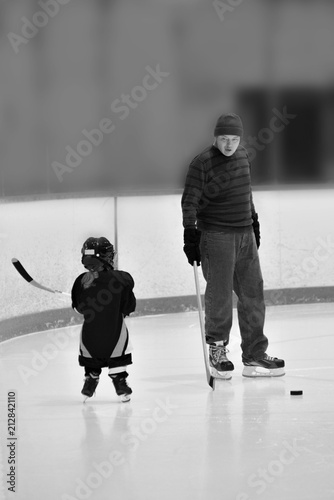 Black and white photo of little hockey girl is wearing in full equipment: helmet, glows, skates, stick. She is holding a stick and looking on a coach. Coach is explaining something