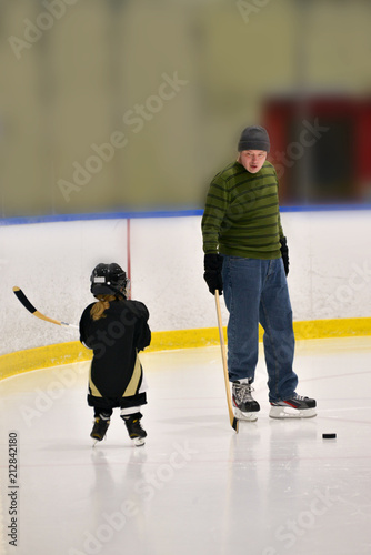 Little hockey girl is wearing in full equipment: helmet, glows, skates, stick. She is holding a stick and looking on a coach. Coach is explaining something