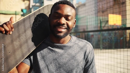 Pleasant-looking African guy in gray T-shirt looking at camera and smiling. Portrait of likable young man with skateboard in hand on background of sports ground.