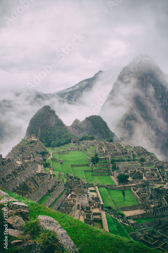 Verical classic view of the ancient mysterious city of Machu Picchu with intense clouds covering the Andes on the background in Peru. Astonishing wallpaper image.
