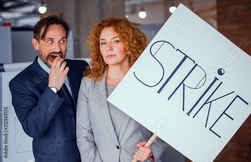 Businesswoman holding big poster with word strike. Red haired fwmale showing unhappy emotions, happy man standing close to her eating cake. Toned concept. photo