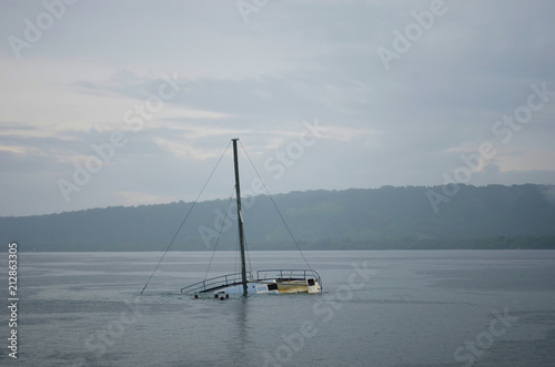 A half-sunken yacht in a misty bay. Its mast rises up against the overcast sky. A tree covered foreshore is in the distance.
