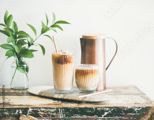 Iced coffee in glasses with milk and straws on board over rustic wooden table, white wall, jug and plant branch in vase at background, horizontal composition. Summer refreshing beverage