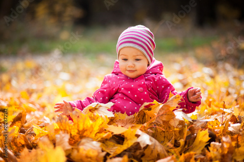 Little kid is playing and sitting in fallen leaves in autumn park. Baby smiles. Girl is dressed in warm hat  jacket.