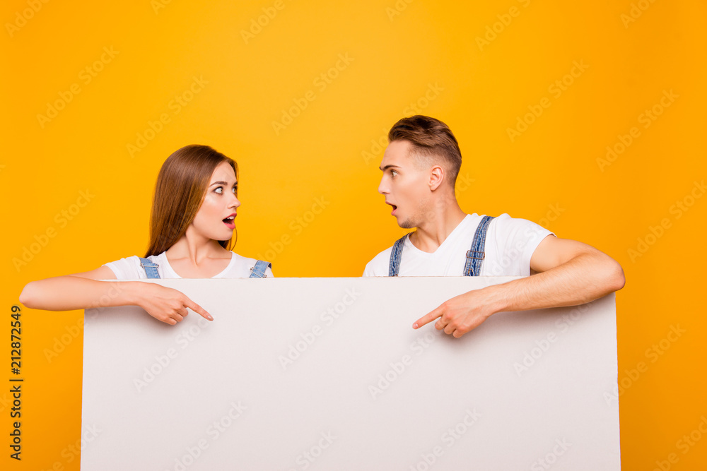 Close up portrait of funny playful lovely beautiful cute couple pointing with fingers to white blank board, showing surprising, looking astonished at each other over yellow background, isolated Close up portrait of funny playful lovely beautiful cute couple pointing with fingers to white blank board, showing surprising, looking astonished at each other over yellow background, isolated