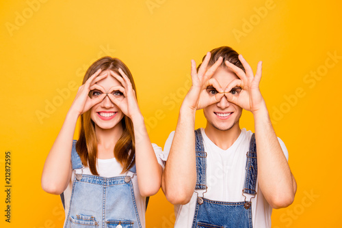 Close up portrait of playful young cute couple wearing casual, showing funny gestures looking through fingers okay sign over yellow background, isolated