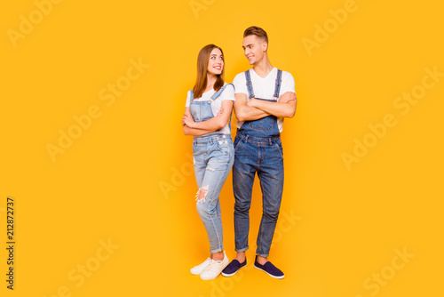 Full lenght portrait of happy lovely adorable young cute couple with folded arms, girl standing back to guy, looking and smiling at each other over yellow background, isolated, copy space © deagreez