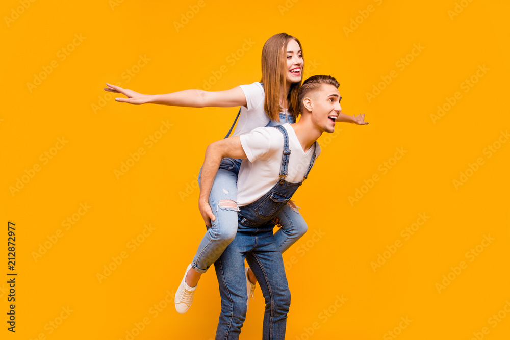 Obraz premium Young funny cheerful couple, boyfriend piggy backing girlfriend, with opened mouths, showing fly gesture with arms wide over yellow background, isolated