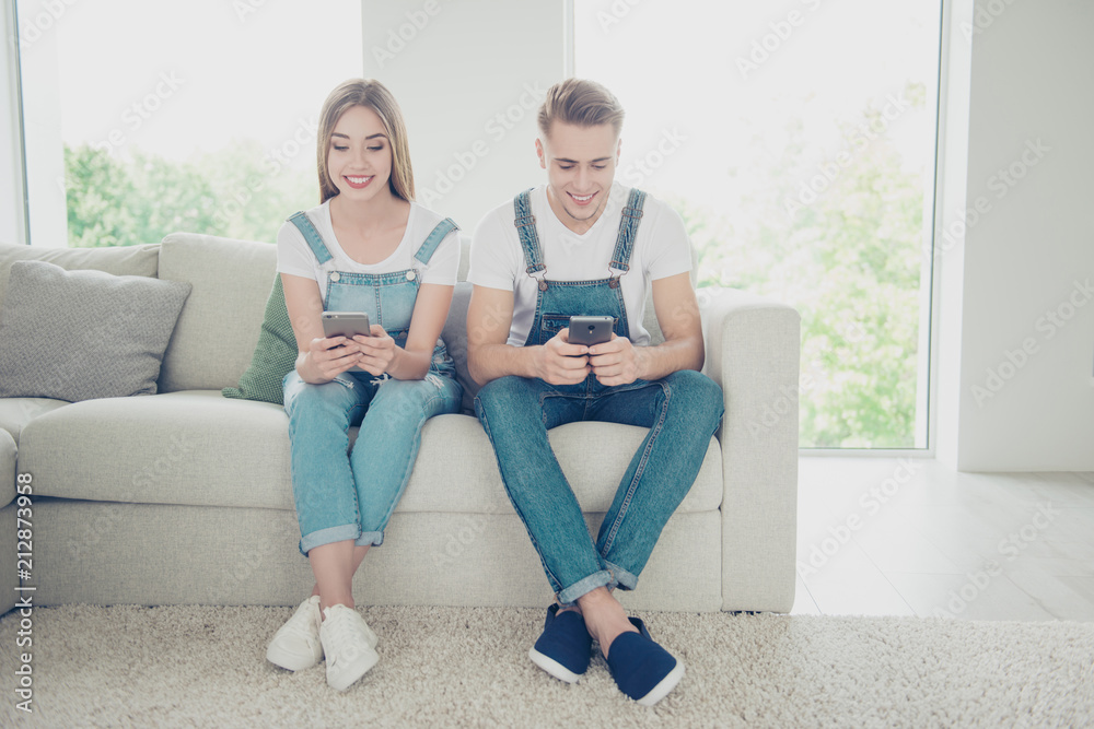 Young attractive beautiful cute couple wearing overall sitting home on sofa and using their smartphones, browsing, reading, surfing the Internet in living room