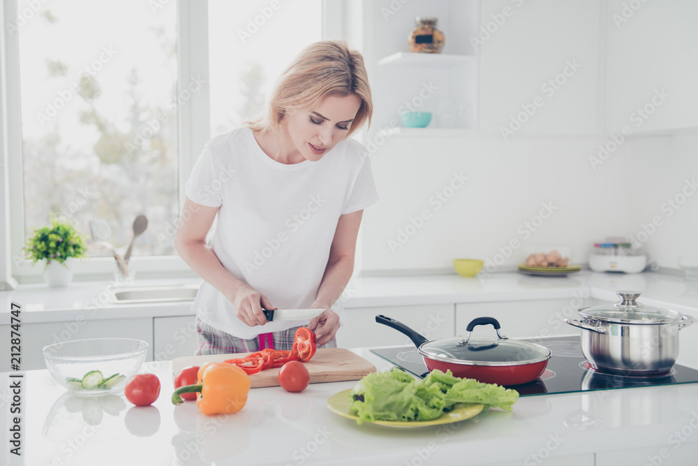 Charming adorable attractive woman wearing pajama chopping with knife red pepper on board in kithcen. Tomatoes, salad, cucumber, pan, pot, yellow pepper lying on table Charming adorable attractive woman wearing pajama chopping with knife red pepper on board in kithcen. Tomatoes, salad, cucumber, pan, pot, yellow pepper lying on table