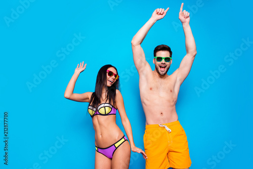 Hot summer time concept. Portrait of cheerful couple in sunglasses and bright swimsuits dance together with beach party mood raise their hands up isolated on bright blue background