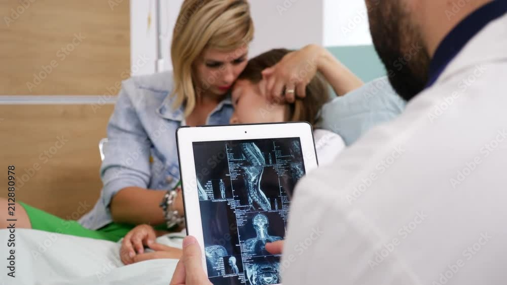Revealing shot of doctor looking on digital tablet PC to MRI scan results while girl patient and her mother are blurred in the background lying on the bed. Revealing shot of doctor looking on digital tablet PC to MRI scan results while girl patient and her mother are blurred in the background lying on the bed.