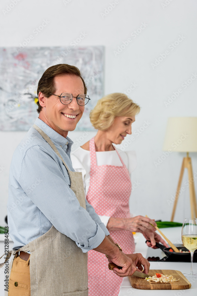 Fototapeta premium side view of grey hair couple cooking vegetables on frying pan and cutting vegetables on wooden board at kitchen