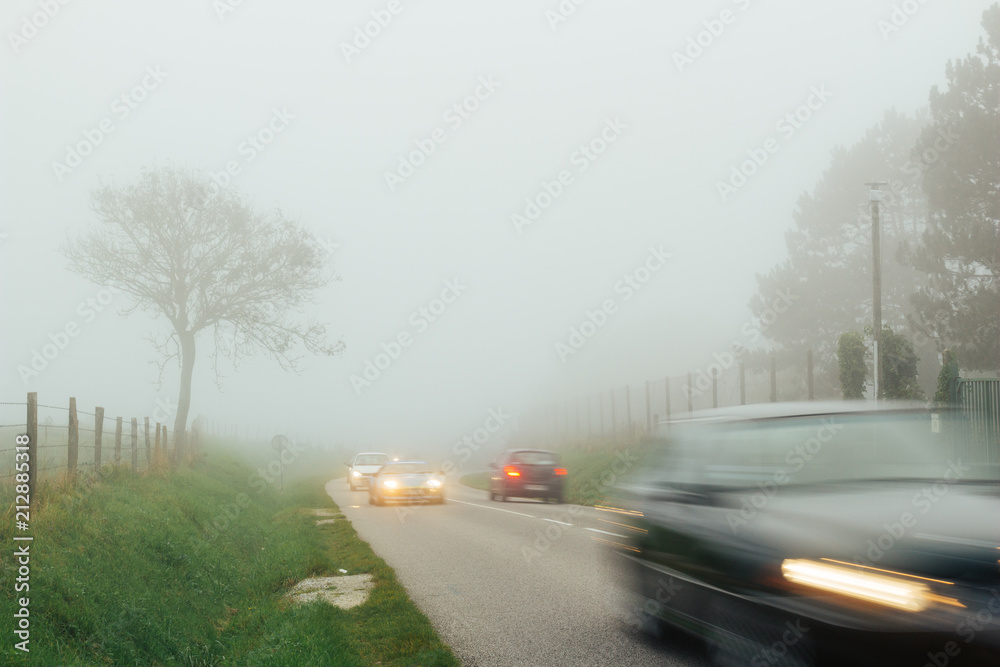 Cars driving on the country asphalt road through the fields on a foggy day in Normandy, France. Twilight countryside landscape, misty weather, car lights. Transport and traffic concept. Long exposure.