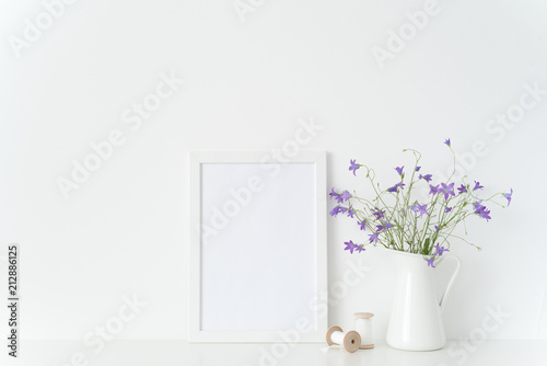 Fototapeta Naklejka Na Ścianę i Meble -  Cute white portrait frame mockup with summtr lilac wild flowers in jug, coils and tapes near wall on white background. Empty frame mock up for presentation design. Template framing for interior art.