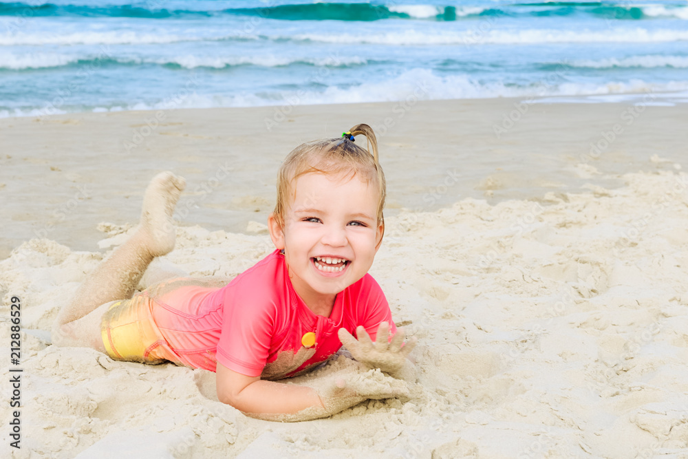 Portrait of emotional adorable toddler girl in sun protecting swimming suit lying on the sand on the beach and looking at camera. Family vacation, traveling concept. Selective focus, copy space. Portrait of emotional adorable toddler girl in sun protecting swimming suit lying on the sand on the beach and looking at camera. Family vacation, traveling concept. Selective focus, copy space.