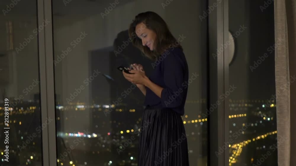 Young woman texting on smartphone standing by window at home at night
Young woman texting on smartphone standing by window at home at night