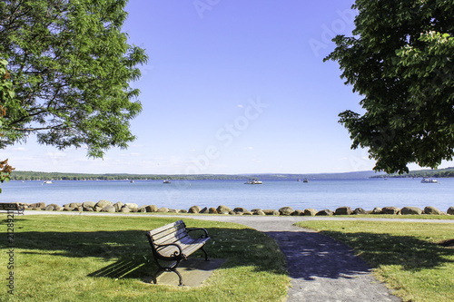 Fototapeta Naklejka Na Ścianę i Meble -  Canandaigua Lake sidewalk through the park. Bench shaded by trees with view of the lake