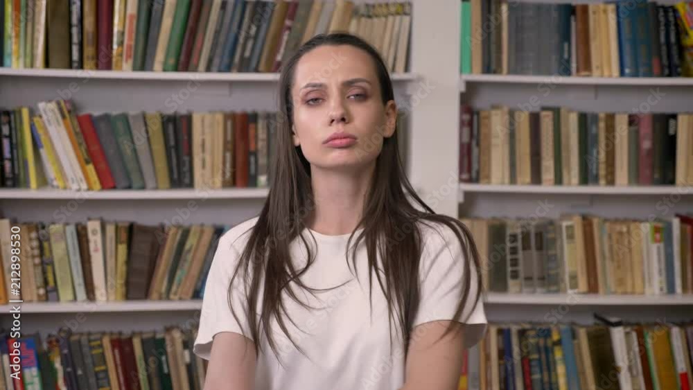 Young serious sexy brunette girl is standing in library, tucking her hair