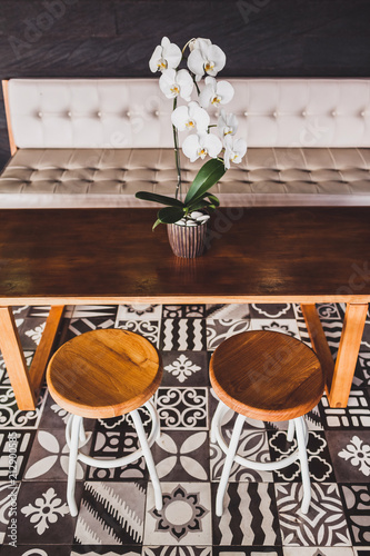 Modern wooden furniture in cafe on floor with black and white ornament tile. Two chairs, table, sofa, orchid flower
