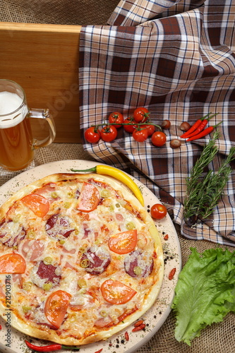 Pizza with tomato, cheese, smoked sausage, boiled sausage, pepper spices on a wooden tray, with beer in a glass mug close-up