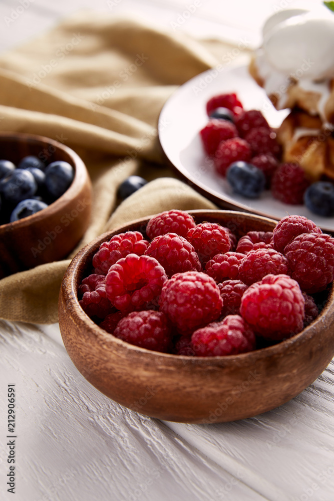 close-up shot of bowls with berries with belgian waffles blurred on background on white wooden table close-up shot of bowls with berries with belgian waffles blurred on background on white wooden table