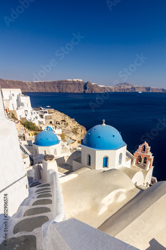 Incredibly romantic scene on Santorini. Fira, Greece. Amazing daytime view towards the deep sea crystal waters with path walk, white houses and blue church roof with white Christian cross and bells