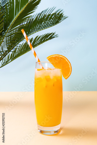 Glass of fresh orange juice and green palm leaves on pastel background. Copy space. Pop art design, minimal summer concept.
