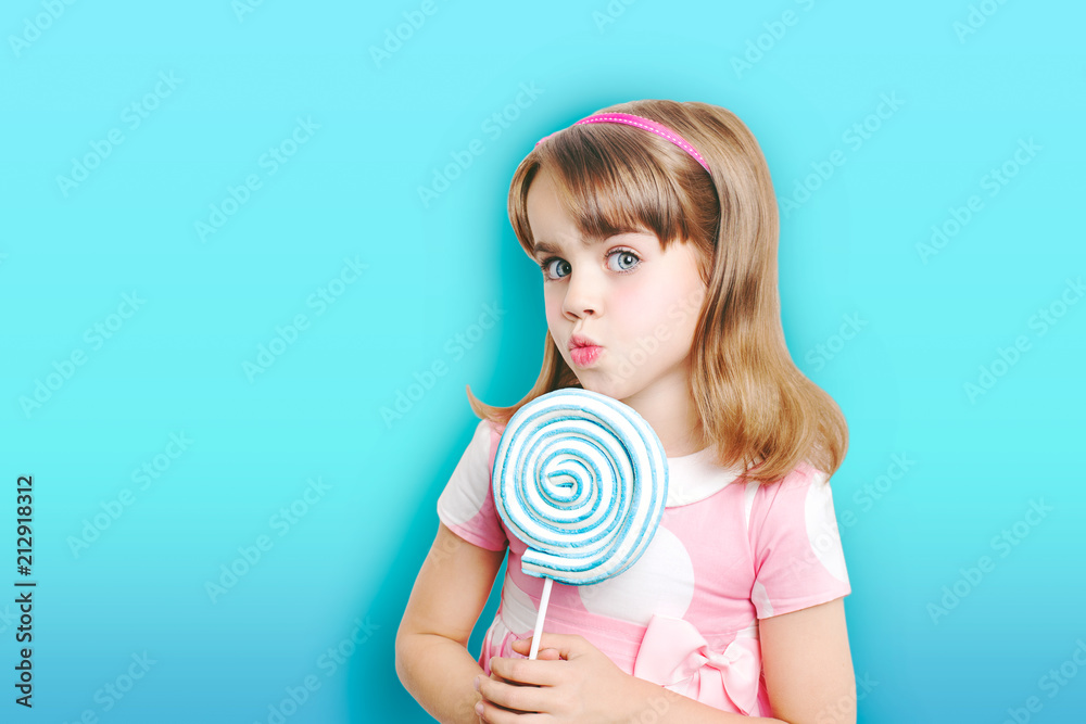 Little girl with lollypop on blue background.Beautiful blond smiling on a light blue  background with a large round striped candy. Attractive girl with lollipop in hand.