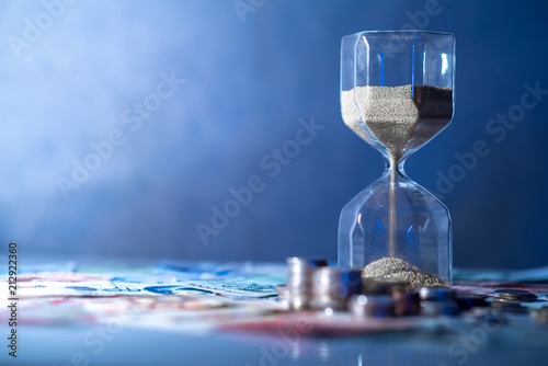 Sand running through the shape of hourglass on table with banknotes and coins of international currency. Time investment and retirement saving. Urgency countdown timer for business deadline concept