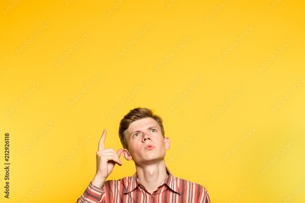 amazed impressed astonished man looking and pointing up with a finger. portrait of a young guy on yellow background popping up or peeking out from the bottom. free space for advertisement. - obrazy, fototapety, plakaty 