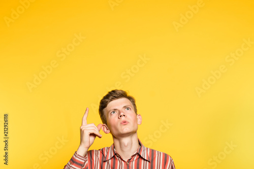 amazed impressed astonished man looking and pointing up with a finger. portrait of a young guy on yellow background popping up or peeking out from the bottom. free space for advertisement. photo
