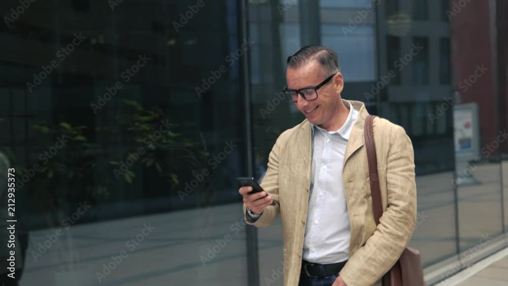 Man in casual wear and stylish glasses walking near modern building and using smartphone