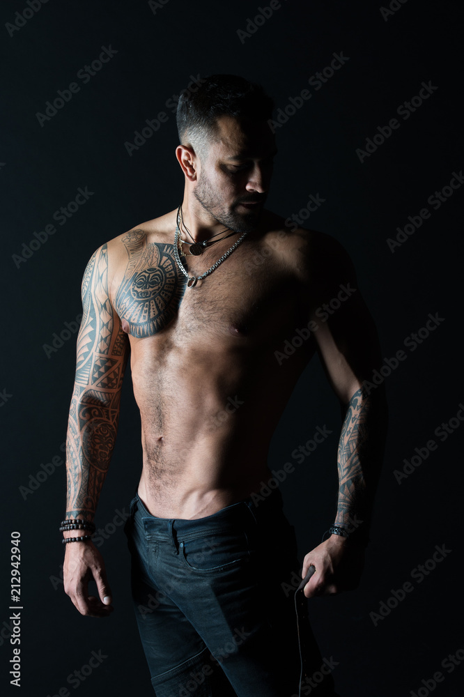 Naklejka premium Bodycare with fitness and sport. Man with tattooed arm and chest. Bearded man with strong torso. Tattoo model with sexy belly. Sportsman or athlete in fashion jeans