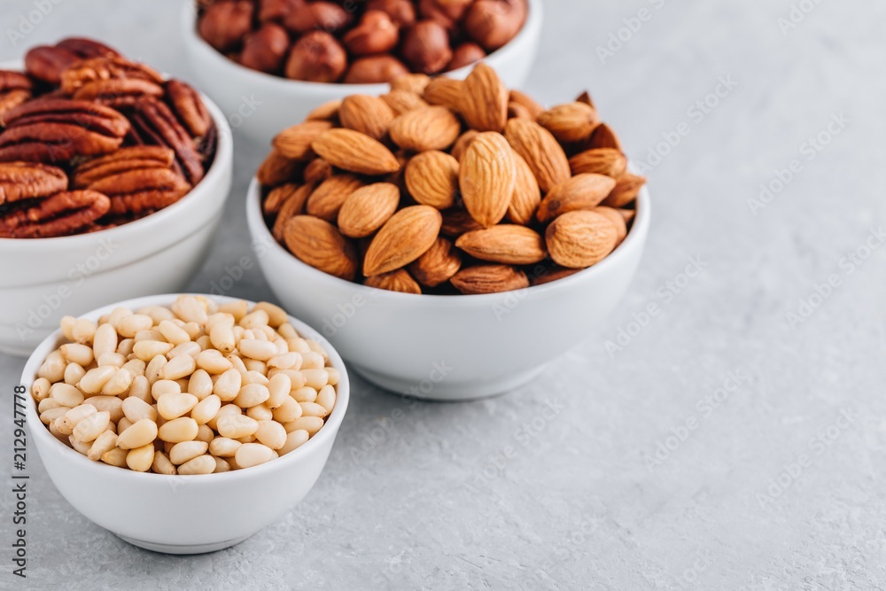 Pine nuts, almonds, pecans and hazelnuts in white bowls on grey background. Mixed nuts. Healthy food and snack. Pine nuts, almonds, pecans and hazelnuts in white bowls on grey background. Mixed nuts. Healthy food and snack.