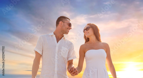 love  summer and relationships concept - happy couple on vacation wearing sunglasses walking and holding hands over sunset sky background