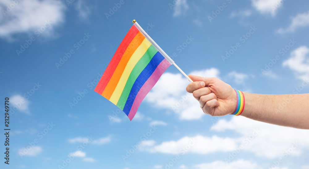 lgbt, same-sex relationships and homosexual concept - close up of male hand with gay pride awareness wristband holding rainbow flag over blue sky and clouds background lgbt, same-sex relationships and homosexual concept - close up of male hand with gay pride awareness wristband holding rainbow flag over blue sky and clouds background