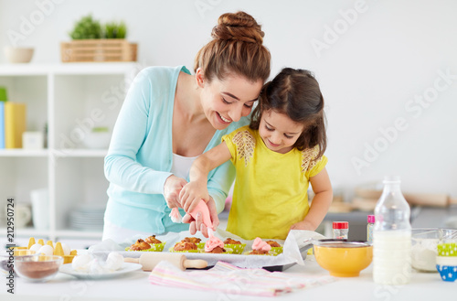 family, cooking and people concept - mother and little daughter making and decorating cupcakes with cream frosting at home kitchen
